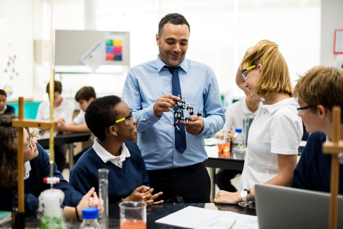 a male teacher with a few students wearing goggles looking at a molecule model